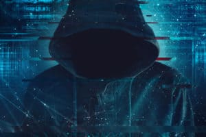 Cybersecurity, computer hacker with hoodie and obscured face, computer code overlaying image. Source; shutterstock.com