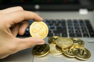 Male businessman hand holding Ripple coin on a background of laptop keyboard and pile of golden coins. Virtual money and Financial growth concept. Trading Mining of Ripples. Source: shutterstock.com