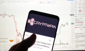 MONTREAL, CANADA - APRIL 26, 2019 Cryptopia cryptocurrency exchange logo and application on Android Samsung Galaxy s9 Plus screen in a hand over a laptop display with bitcoin chart on it. - Image